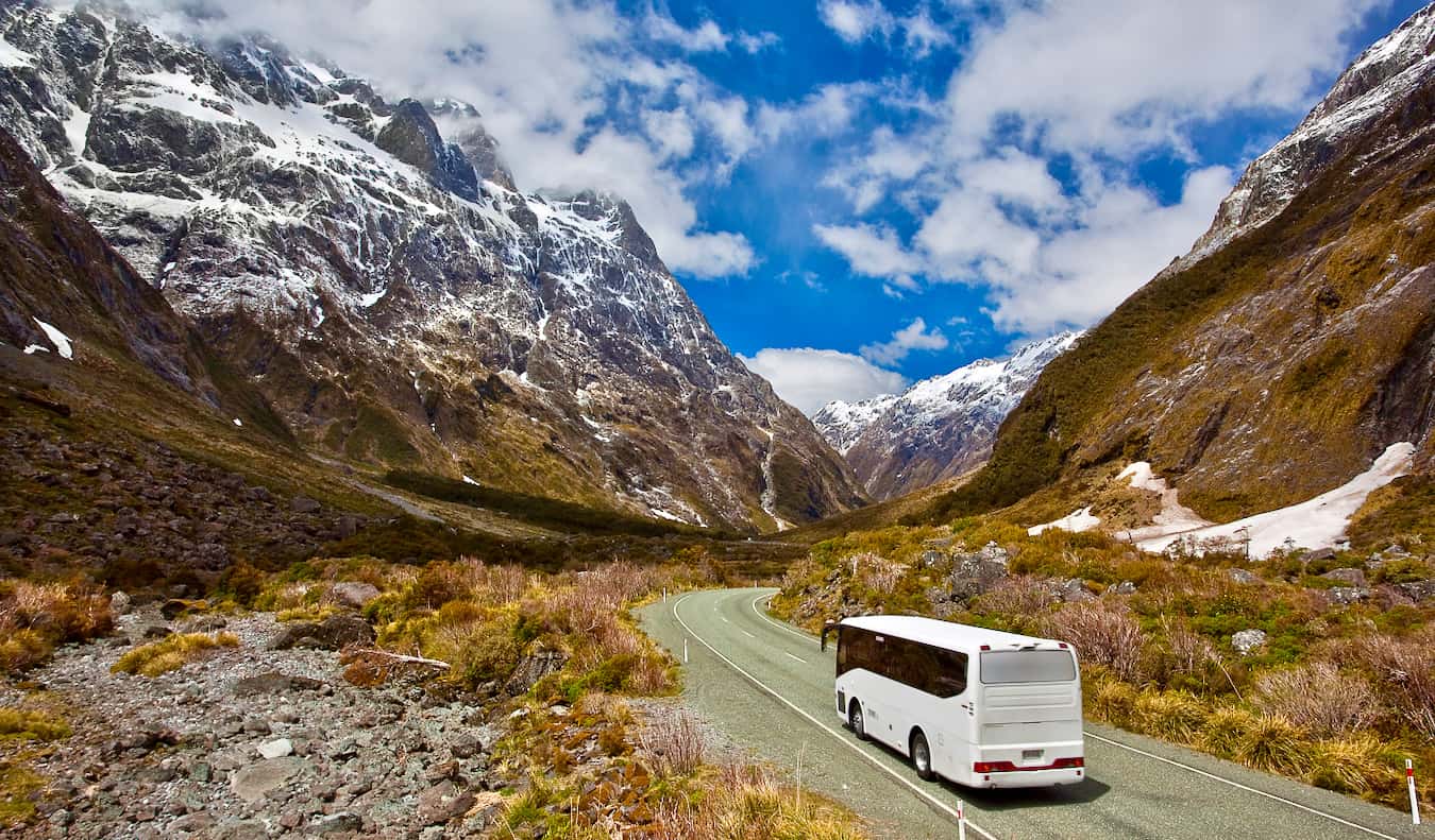 A bus driving down a winding road through tall snowcapped mountains in New Zealand